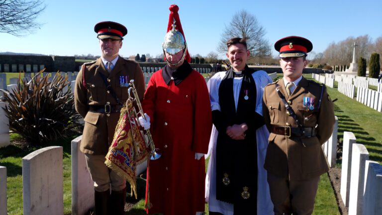 Four men in military uniforms and ceremonial attire stand together at a cemetery honoring a missing Windsor soldier from World War One, with rows of white headstones solemnly stretching into the background.
