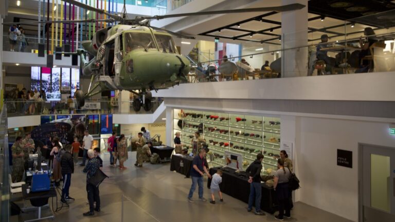 The museum interior comes alive with a spotlight illuminating a helicopter suspended from the ceiling. Visitors, some in military uniforms, observe a meticulously curated exhibit of military artifacts, including pieces from the distinguished Royal Engineers.