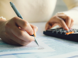 A person in a white sweater diligently fills out forms, possibly related to Contributions Relief, using a pen while navigating numbers on a calculator amidst a desk cluttered with papers.