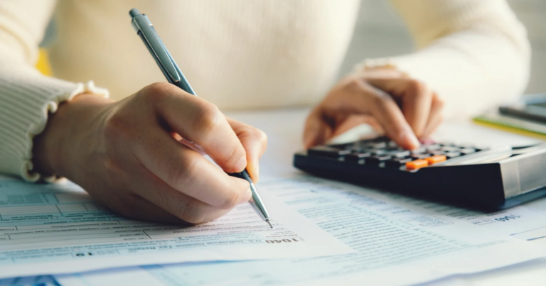 A person in a white sweater diligently fills out forms, possibly related to Contributions Relief, using a pen while navigating numbers on a calculator amidst a desk cluttered with papers.