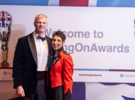 A man in a tuxedo and a woman in a red military-style jacket pose in front of a "Welcome to #SoldieringOnAwards" backdrop with British-themed statues on each side.