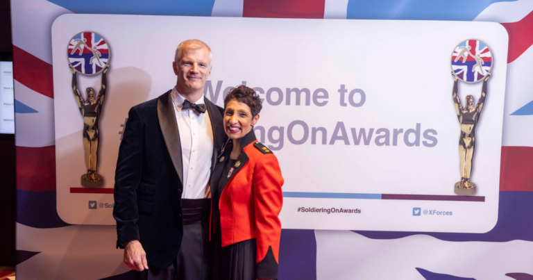 A man in a tuxedo and a woman in a red military-style jacket pose in front of a "Welcome to #SoldieringOnAwards" backdrop with British-themed statues on each side.