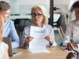A former servicewoman with glasses discusses a document featuring a bar graph during a meeting with four colleagues seated around the table, drawing from her experience transitioning from the military.
