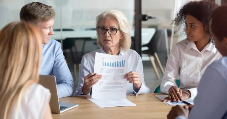 A former servicewoman with glasses discusses a document featuring a bar graph during a meeting with four colleagues seated around the table, drawing from her experience transitioning from the military.