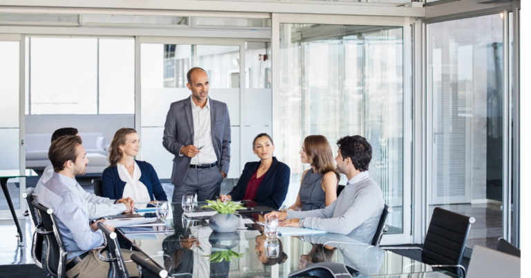 A group of professionals in business attire sits around a conference table in a glass-walled meeting room, attentively listening to a speaker providing an update on the Revisited Team's latest initiatives.