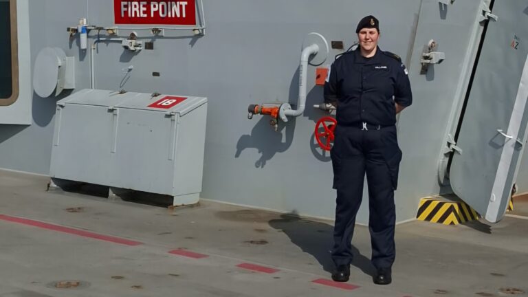 A person in a navy uniform stands on the deck of a ship, by your side next to a fire point sign and a red valve.