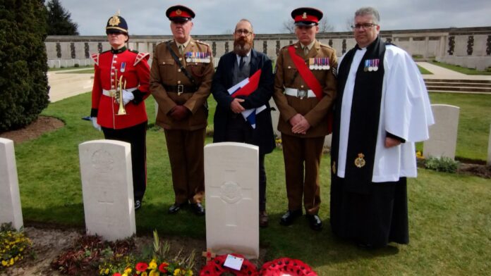 A group of five men, including military personnel in uniform and a clergyman, stand solemnly at a gravesite with wreaths and flowers. The cemetery's wall of inscribed names honors the Unknown Soldiers found from World War One.