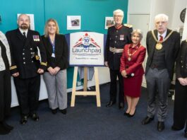 Seven people, including four in formal attire with medals, stand beside a sign for a veterans' support organization dedicated to helping homeless veterans. The backdrop is an office setting in Liverpool, adorned with photographs on the wall.