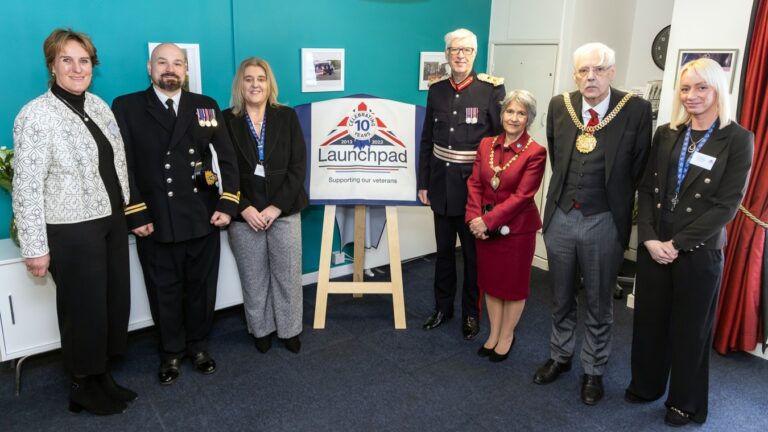 Seven people, including four in formal attire with medals, stand beside a sign for a veterans' support organization dedicated to helping homeless veterans. The backdrop is an office setting in Liverpool, adorned with photographs on the wall.