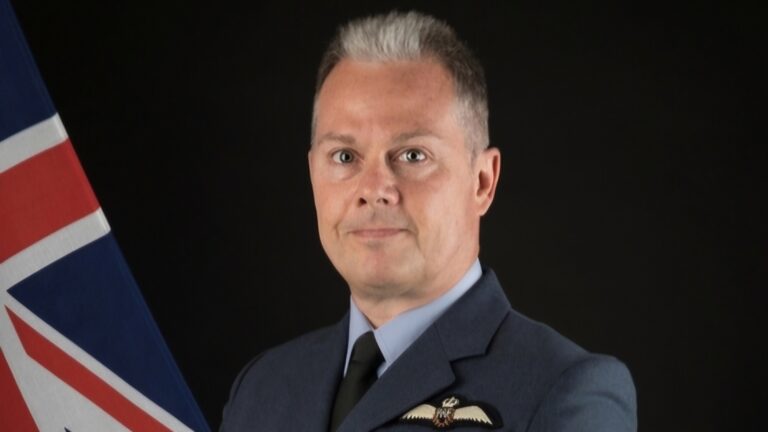 A person in a military uniform poses beside a flag against a dark background, embodying the dedication and commitment SSAFA supports, as emphasized by their CEO.