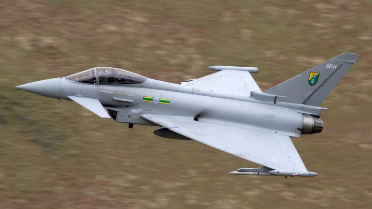 A grey military jet aircraft, specifically a Eurofighter Typhoon, flies at low altitude over a blurred landscape background—a prime example of UK Fighters patrolling NATO airspace.