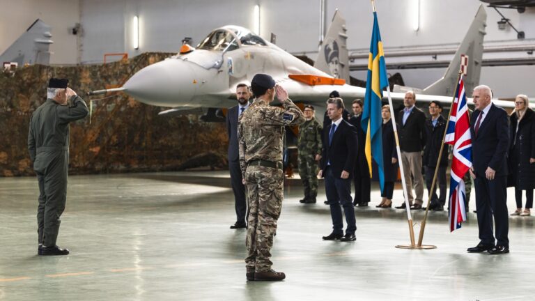 A military ceremony unfolds in a hangar as RAF personnel salute near the British and Swedish flags, with a fighter jet poised to guard NATO's eastern flank.