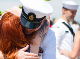 A person in a navy uniform hugs their partner with red hair while holding a rose; other Tri Service members in similar uniforms are seen in the background.