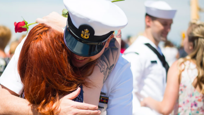 A person in a navy uniform hugs their partner with red hair while holding a rose; other Tri Service members in similar uniforms are seen in the background.