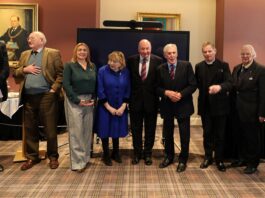 Eight adults in business attire stand side by side indoors in front of a large screen, with framed portraits behind them, following a military fundraiser attended by the Chief of the General Staff.