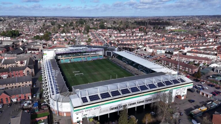 Aerial view of a large stadium surrounded by residential houses. The stadium has a green field and solar panels on the roof. The surrounding area is densely populated with buildings and roads.