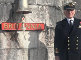 An older man in a naval uniform stands next to a ship's bell and a sign reading "Britannia" mounted on a stone wall, capturing a true full circle moment in maritime tradition.