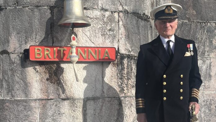 An older man in a naval uniform stands next to a ship's bell and a sign reading 