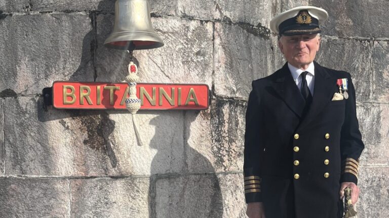 An older man in a naval uniform stands next to a ship's bell and a sign reading "Britannia" mounted on a stone wall, capturing a true full circle moment in maritime tradition.