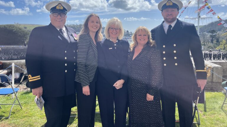 Five people pose outdoors, with two men in naval uniforms on either end—symbols of dedicated service—and three women in civilian clothing in the center, all set against a ship and cloudy sky, reflecting pride in their naval community.