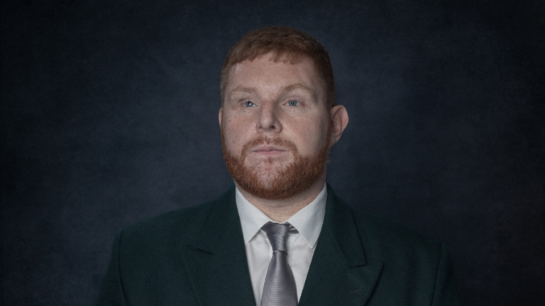A man with short red hair and a full beard, wearing a dark suit, white shirt, and silver tie, stands against a dark, textured background—embodying the quiet confidence of everyday heroes shaping the future.