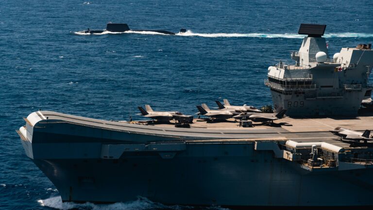 A UK Carrier Strike Group aircraft carrier with fighter jets on its deck sails in the Mediterranean, with a submarine partially visible in the water nearby.