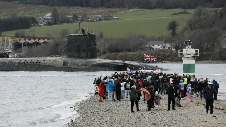 A crowd of people, united in support, gather on a rocky shoreline to watch a British-flagged submarine sail by under a cloudy sky. Among them are submariner families, proudly awarded recognition for their loved ones' service and dedication.