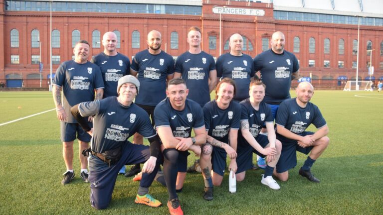 A group of eleven men in navy blue soccer uniforms pose for a team photo on a sports field in front of a red brick building labeled "RANGERS F.C.," showcasing their Rangers FC spirit and strong Football Partnership.