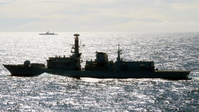 A British warship sails on a calm, sunlit sea with another ship visible in the distant background, patrolling UK waters.