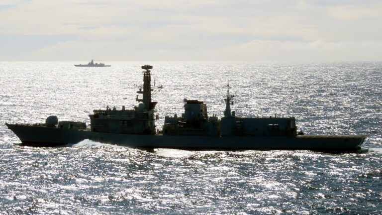 A British warship sails on a calm, sunlit sea with another ship visible in the distant background, patrolling UK waters.