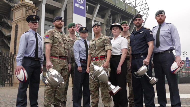 A group of military personnel in various uniforms stand holding trophies and rugby balls in front of a stadium, celebrating the thrilling Army v Navy Rugby match. JJ Chalmers, the live host, shares a proud smile with the victors.