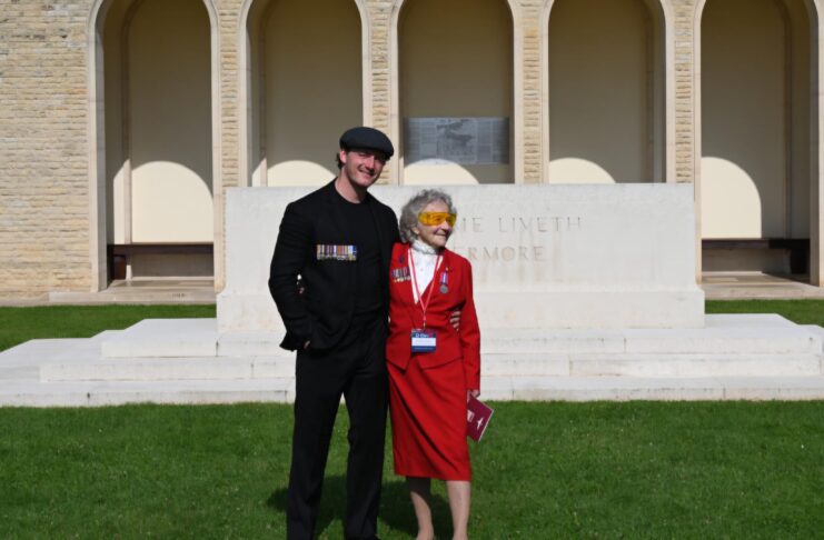 A man and an elderly woman, both supported by the Taxi Charity, stand together before a war memorial. The woman wears a red outfit adorned with medals, while the man is dressed in black with a flat cap—honoring our heroes.