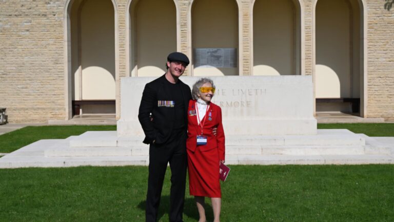 A man and an elderly woman, both supported by the Taxi Charity, stand together before a war memorial. The woman wears a red outfit adorned with medals, while the man is dressed in black with a flat cap—honoring our heroes.