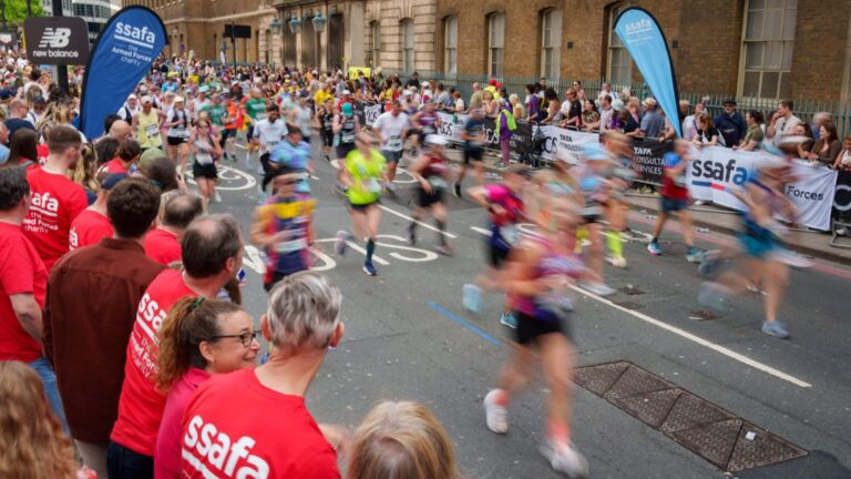 A crowd of spectators in red shirts cheers on a group of marathon runners during the London Marathon, with banners and supporters rallying for a Military Charity lining the city street.