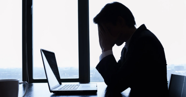A silhouetted male veteran sits at a desk, overwhelmed by challenging experiences, holding his head in his hands in front of an open laptop, with a large window in the background.