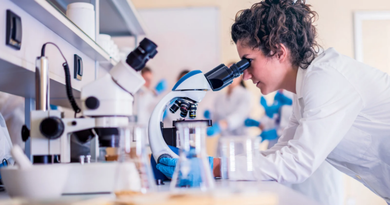 A scientist in a lab coat looks through a microscope at a laboratory bench, surrounded by glassware and equipment—an environment where medical research funding drives every material change and breakthrough.