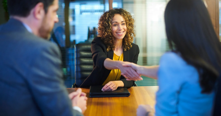 A woman in business attire smiles and shakes hands with another person across a table, while a man sits beside them in an office setting, highlighting positive connections between employers and job seekers.