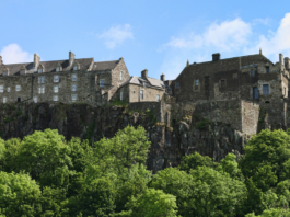 Recognised as a key landmark for Stirling 900, this stone castle complex sits atop a rocky hill, surrounded by dense green trees under a blue sky with scattered clouds.