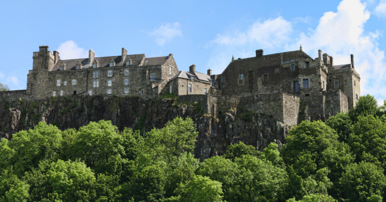 Recognised as a key landmark for Stirling 900, this stone castle complex sits atop a rocky hill, surrounded by dense green trees under a blue sky with scattered clouds.