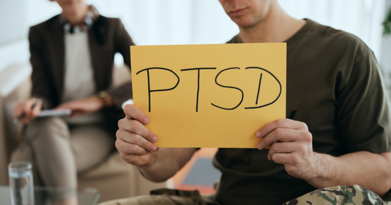 A person in a military-style shirt holds a yellow "PTSD" sign, highlighting veterans mental health; another individual sits in the background, possibly in a therapy setting.