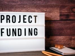 A lightbox sign displays the words "Project Funding" next to a notepad, pencil, and wooden stamp on a wooden surface—highlighting support for Seldom Heard Needs within Armed Forces Communities.