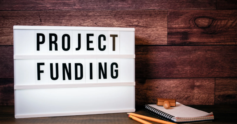 A lightbox sign displays the words "Project Funding" next to a notepad, pencil, and wooden stamp on a wooden surface—highlighting support for Seldom Heard Needs within Armed Forces Communities.