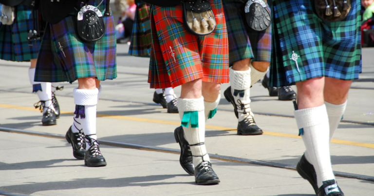 A group of people wearing kilts, knee-high socks, and formal shoes march in a parade on a paved street to raise awareness for veterans’ mental health.