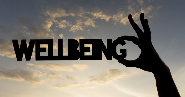 A silhouetted hand makes an "OK" gesture, framing the word "WELLBEING" against a cloudy sunset sky—capturing the spirit of Creative Wellbeing and supporting the Armed Forces Community.