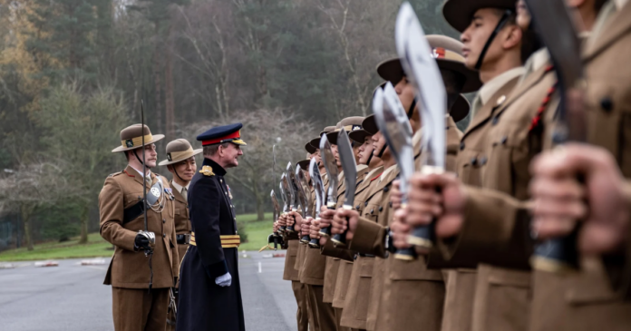 A military officer inspects a row of uniformed soldiers holding kukri knives, with trees and overcast sky in the background.