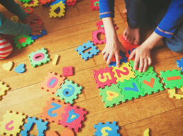 Two children play on a wooden floor with colorful foam puzzle pieces featuring numbers and shapes.