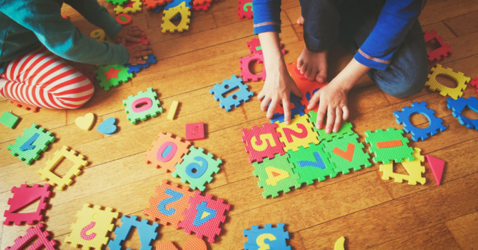 Two children play on a wooden floor with colorful foam puzzle pieces featuring numbers and shapes.