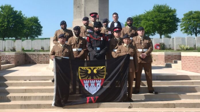A group of uniformed soldiers, possibly Unknown Soldiers buried in France, pose on steps before a monument, holding a black flag with a crest and the Roman numeral IV.