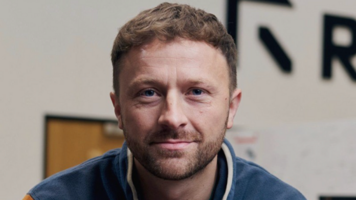 A man with short brown hair and beard, wearing a blue jacket, looks directly at the camera indoors with a blurred background—he could easily be mistaken for a tech firm professional.