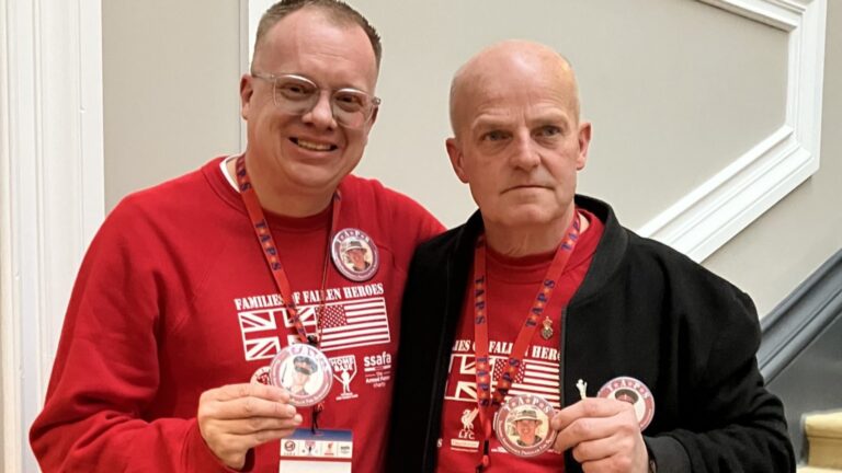 Two men wearing red tops with printed designs stand side by side, holding circular badges featuring a photo. In an indoor setting with white paneled walls, their expressions reflect a shared journey, reminiscent of veterans honoring memories beyond the football field.